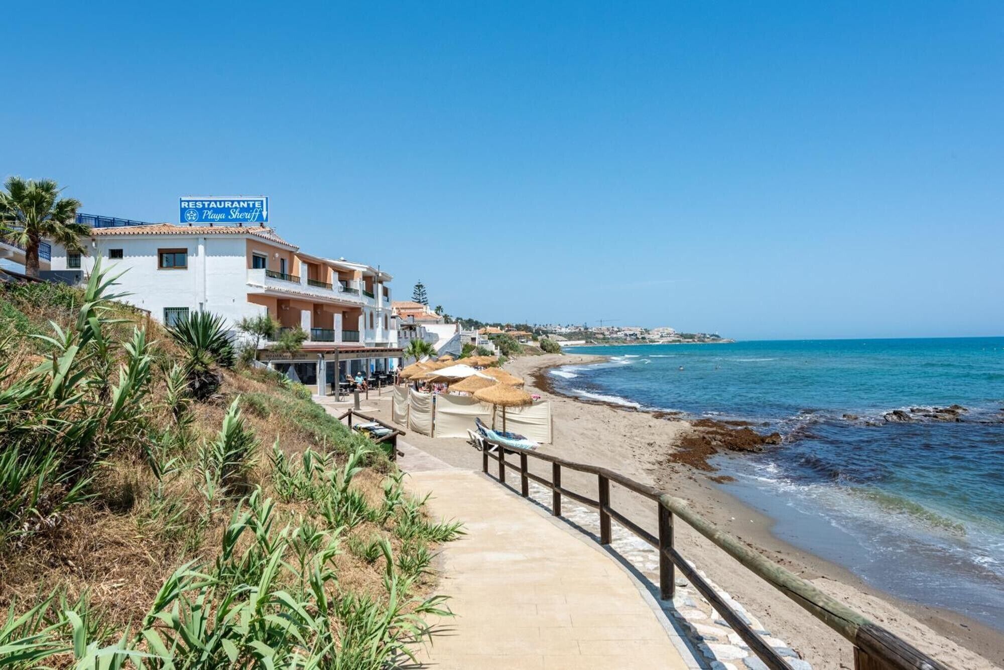 A coastal hotel with a paved walkway leading to the beach, a sign for 'Restaurante Playa Sheriff,' and a view of the sea.