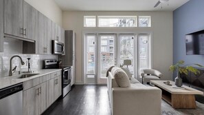 Une cuisine moderne avec des meubles gris, un four à micro-ondes et un lave-vaisselle. On y trouve une grande fenêtre avec des stores, un canapé et une table basse.