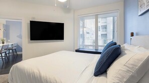 Ein modernes Hotelzimmer mit einem großen Bett, einem flachbildfernseher an der Wand, einem kleinen Schreibtisch mit Stuhl und einem Fenster mit Jalousien.