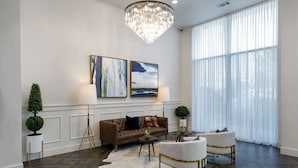 Un salon moderne avec un canapé en cuir marron, deux fauteuils blancs, un lustre et de grandes fenêtres ornées de rideaux légers.