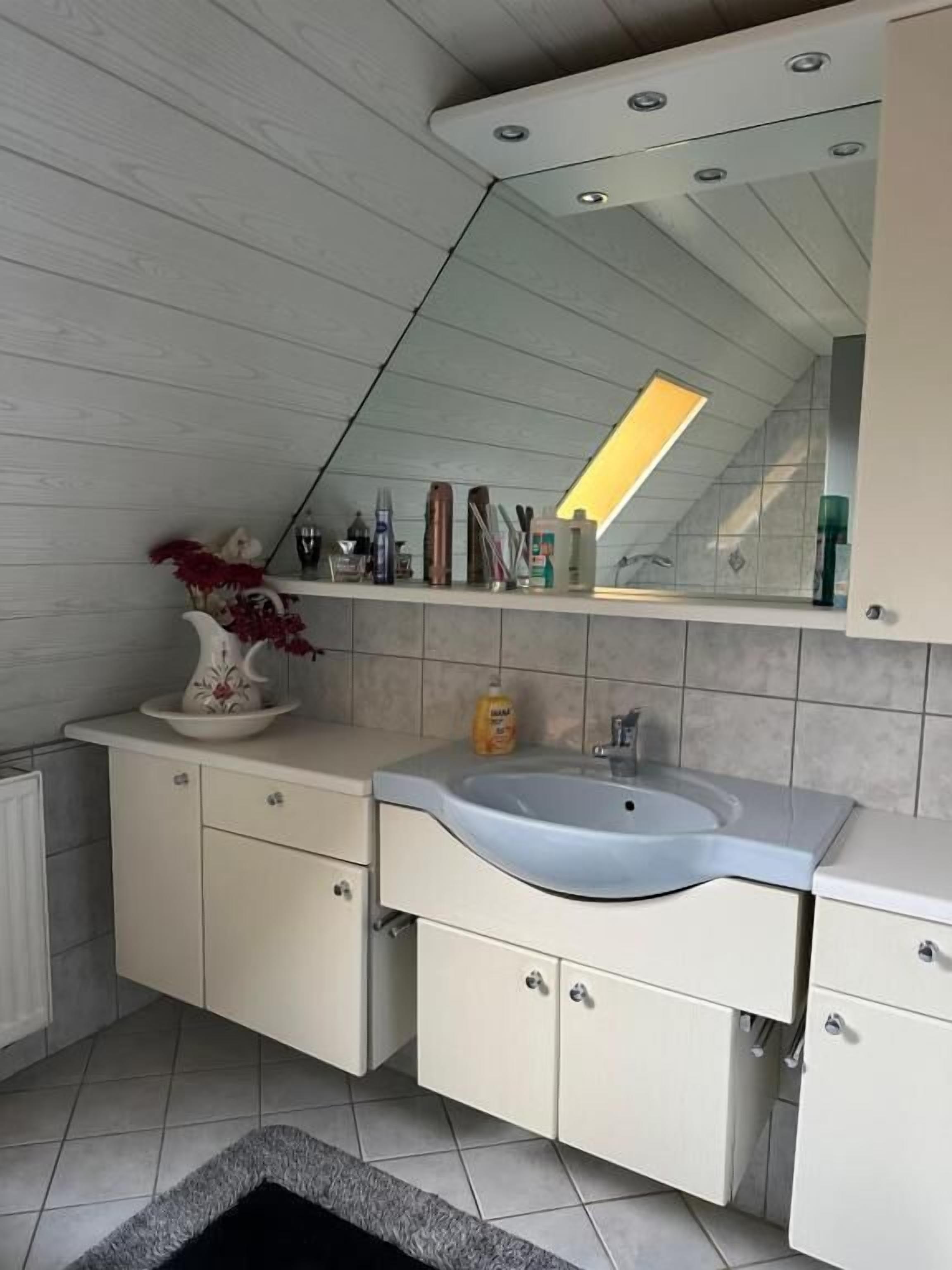 Een moderne badkamer met een witte wastafel, een wastafel en een spiegel erboven. Op de wastafel staat een bloemenvaas en er liggen toiletartikelen op het aanrecht.