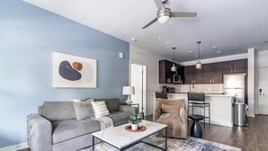 Una sala de estar moderna con un sofá gris, una mesa de centro y un tapete azul y blanco. Se ve una cocina al fondo con armarios oscuros y electrodomésticos de acero inoxidable.