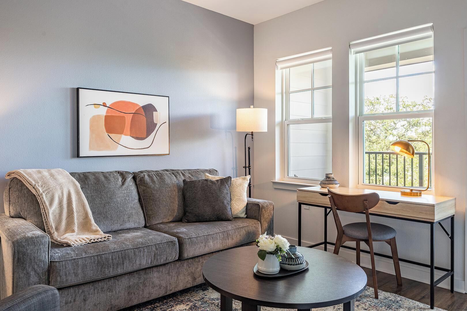 A modern living room with a gray sofa, a coffee table with decorative items, a desk with a chair, and a large window with a view of greenery.