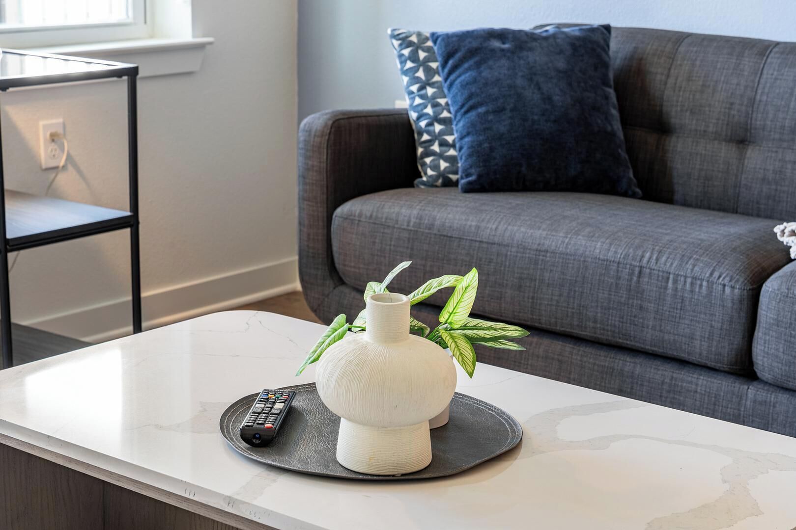 Un salón moderno con un sofá gris, un jarrón blanco con hojas verdes y un control remoto sobre una mesa blanca.