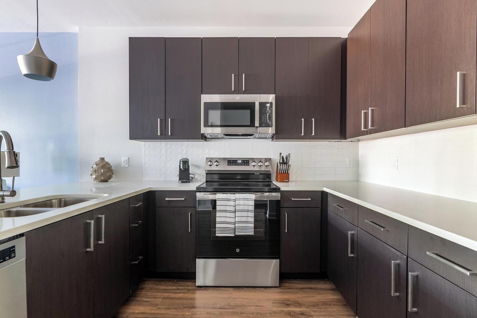A modern kitchen with dark cabinets, stainless steel appliances, and a wooden floor.