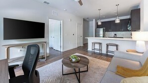 Una sala de estar moderna con una televisión de pantalla plana, un escritorio con una silla, un sofá, una mesa de centro con una planta, un ventilador de techo y una zona de cocina con taburetes de bar.