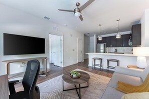 Ein modernes Wohnzimmer mit Flachbildfernseher, Schreibtisch mit Stuhl, Sofa, Couchtisch mit Pflanze, Deckenventilator und einer Küchenzeile mit Barhockern.