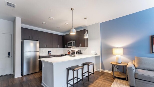 Una cocina moderna con una isla central, armarios oscuros, electrodomésticos de acero inoxidable, iluminación colgante y una zona de estar acogedora con una lámpara.