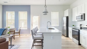 A modern kitchen with a central island, stainless steel appliances, and a dining area with a sofa and chairs. - Landing | Stunning 3BD, Gym, Yoga Studio (Orange Beach)