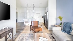 A modern living room with a sofa, a coffee table, and a TV. There is a kitchen area with white cabinets and a refrigerator. - Landing | Amazing 1BD, Gym, Pool (Orange Beach)