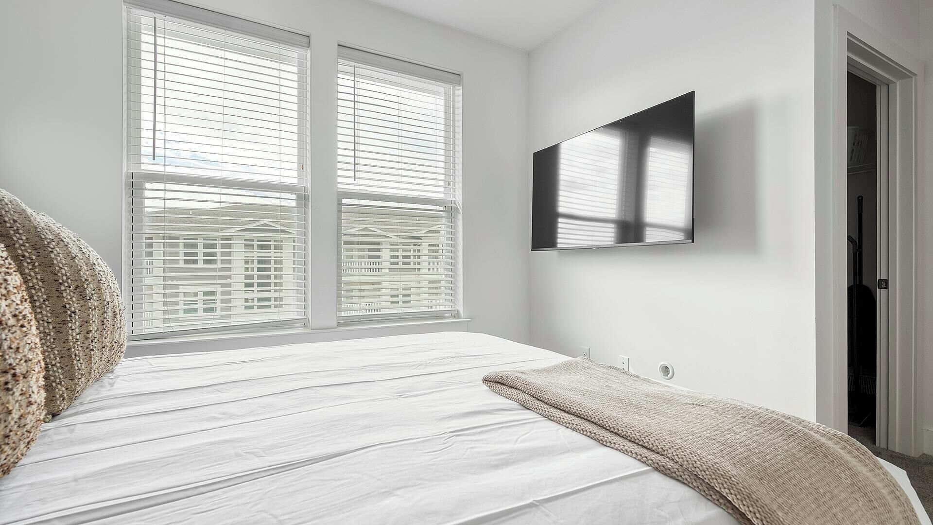 A bedroom with a large bed, two windows with blinds, a flat-screen TV mounted on the wall, and a door to the right.
