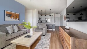 Una sala de estar moderna con un sofá gris, una mesa de centro y una planta. Hay una zona de cocina con armarios blancos y una encimera de madera.