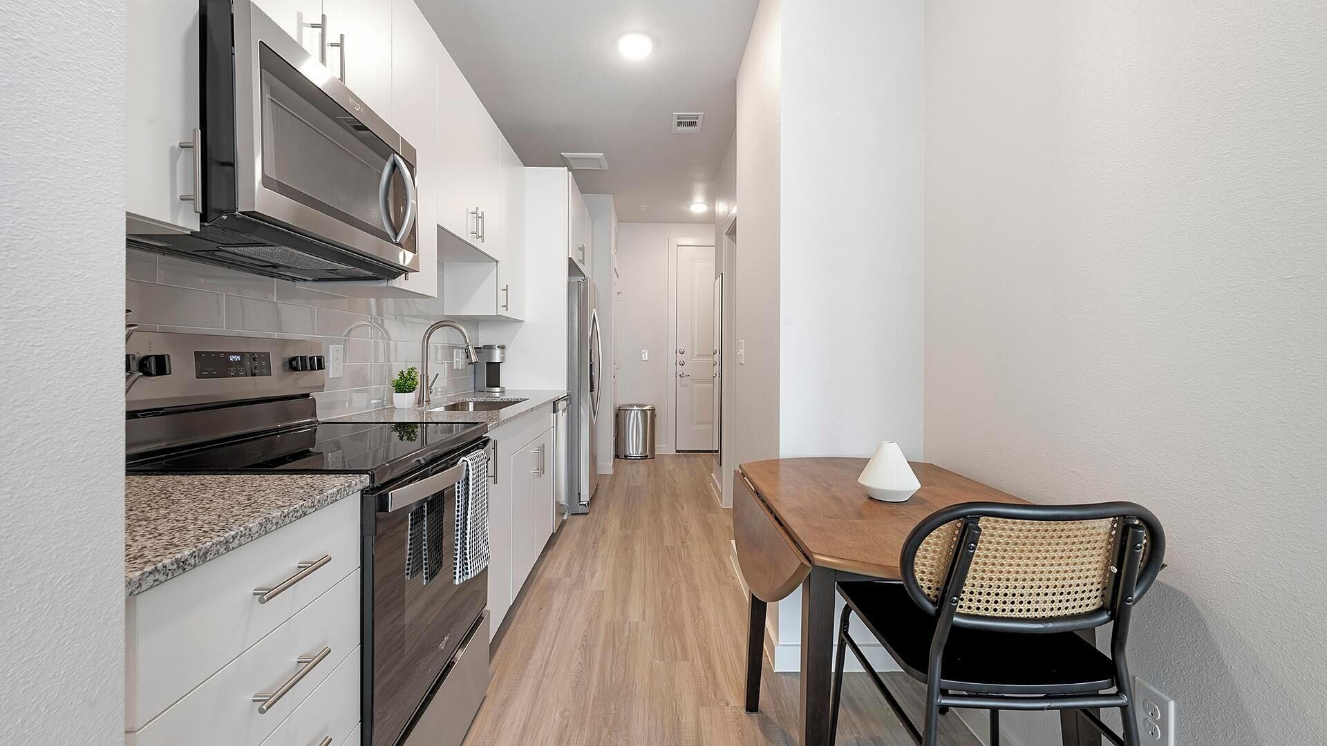 A compact kitchen with modern appliances, a dining area with a wooden table and a chair, and a hallway leading to a door.