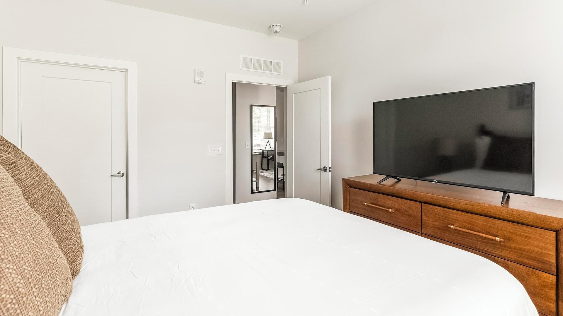 A bedroom with a bed, a wooden dresser, a TV, and a door leading to a bathroom.