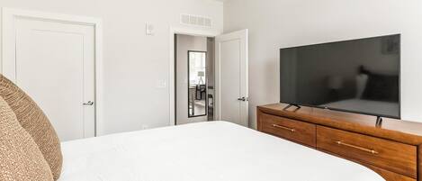 A bedroom with a bed, a wooden dresser, a TV, and a door leading to a bathroom.