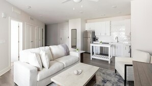A modern living room with a white sofa, a coffee table, and a kitchen area with white cabinets and a refrigerator. - Landing | Incredible 1BD, Clubhouse, Gym (Huntsville)