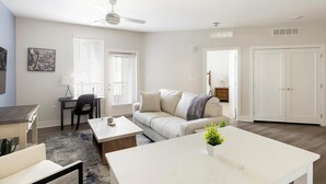 A modern living room with a sofa, coffee table, and a dining table. There is a desk with a chair, a ceiling fan, and a door leading to another room. - Landing | Incredible 1BD, Clubhouse, Gym (Huntsville)