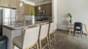 Uma cozinha moderna com frigorífico de aço inoxidável, micro-ondas e fogão. Há dois bancos altos e uma pequena mesa com uma cadeira.
