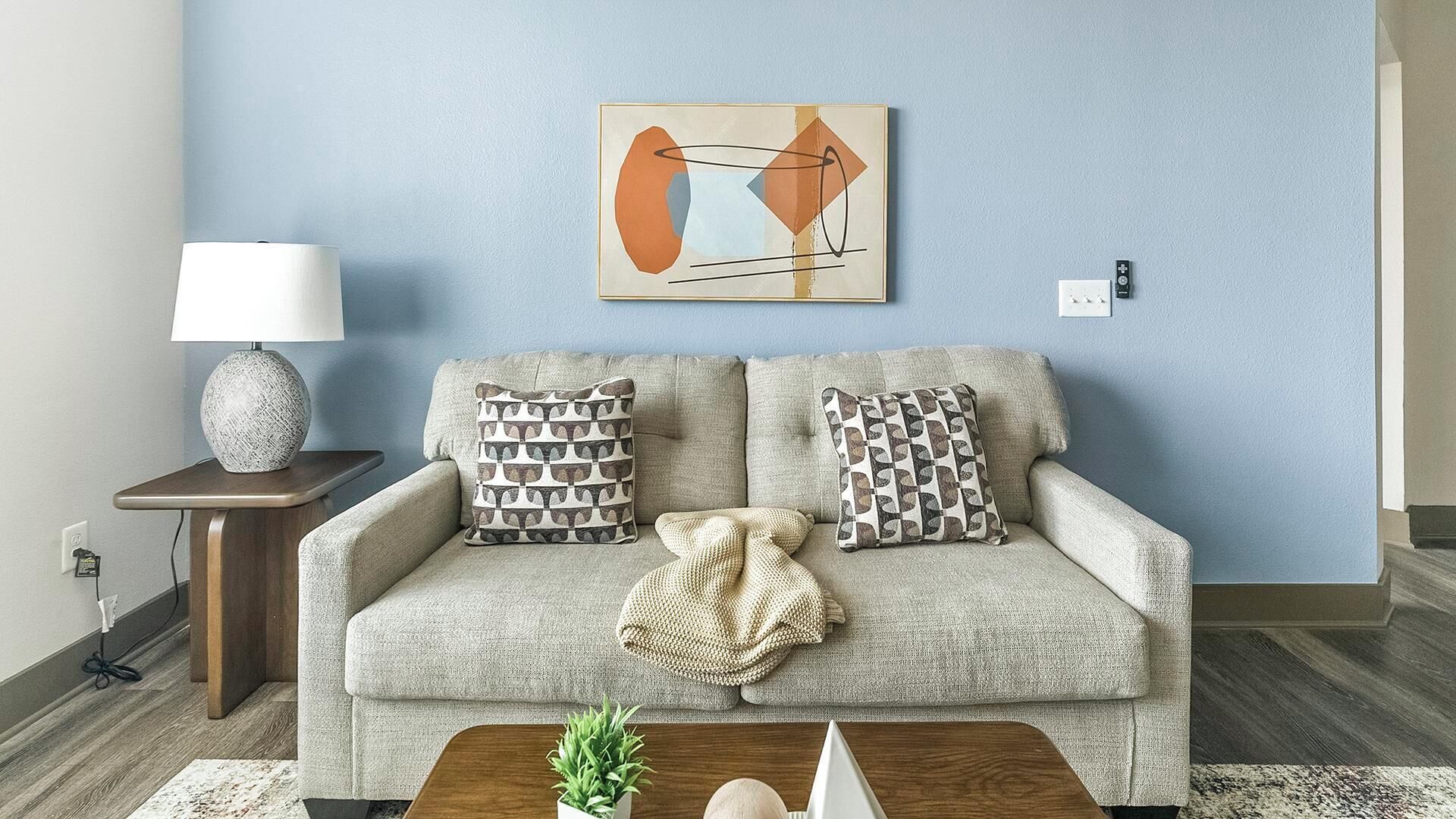 Una sala de estar con un sofá, cojines estampados, una manta, una lámpara y un cuadro en la pared.