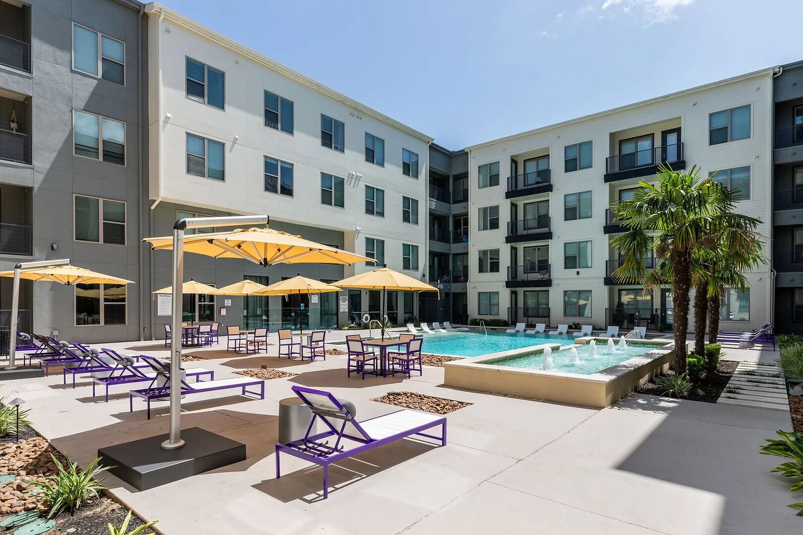 A modern outdoor pool area with lounge chairs and umbrellas, surrounded by a multi-story building with balconies.