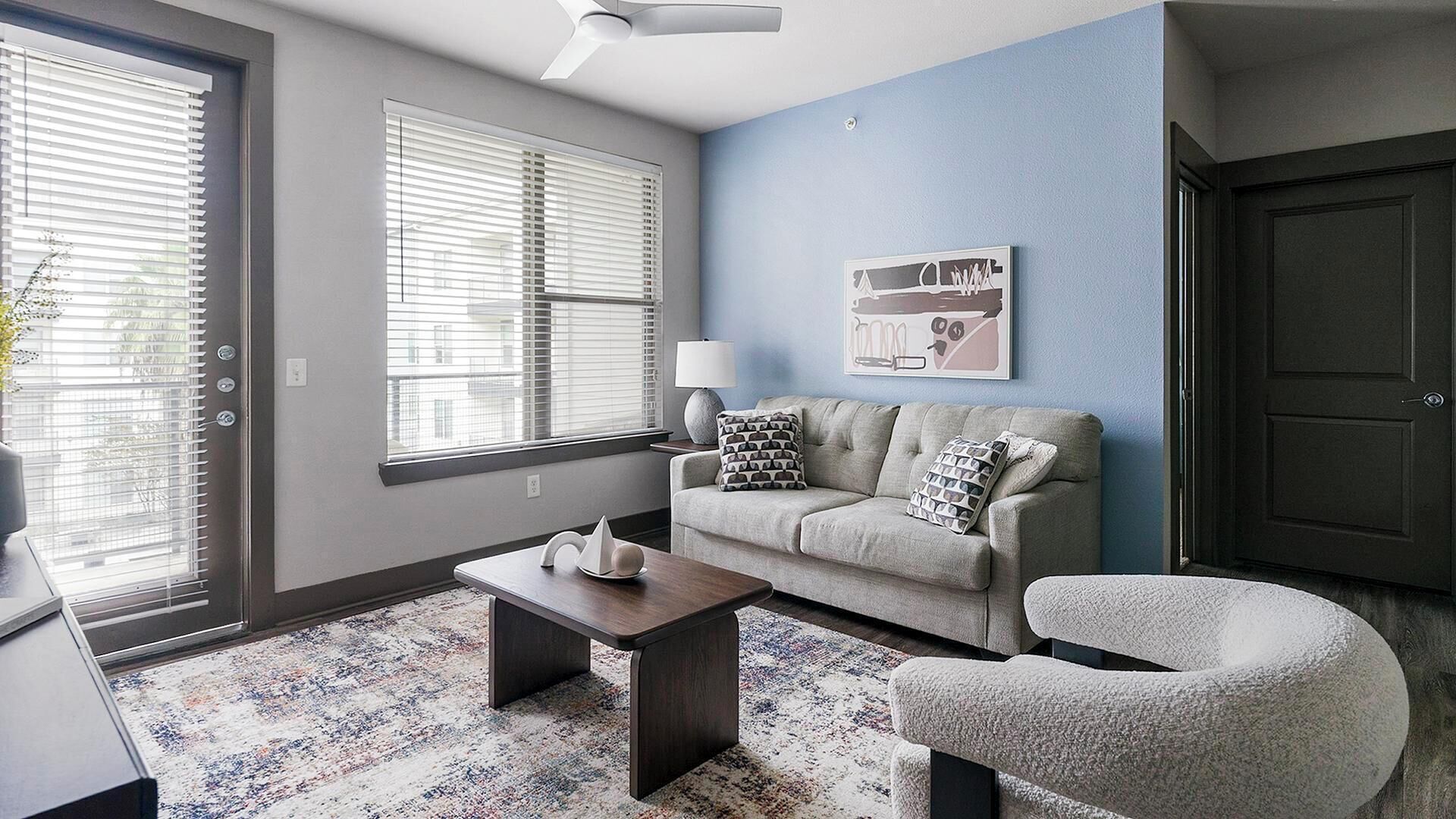 A living room with a sofa, armchair, coffee table, and a lamp. There are two windows with blinds, a door, and a wall-mounted artwork.