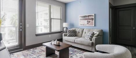 A living room with a sofa, armchair, coffee table, and a lamp. There are two windows with blinds, a door, and a wall-mounted artwork.