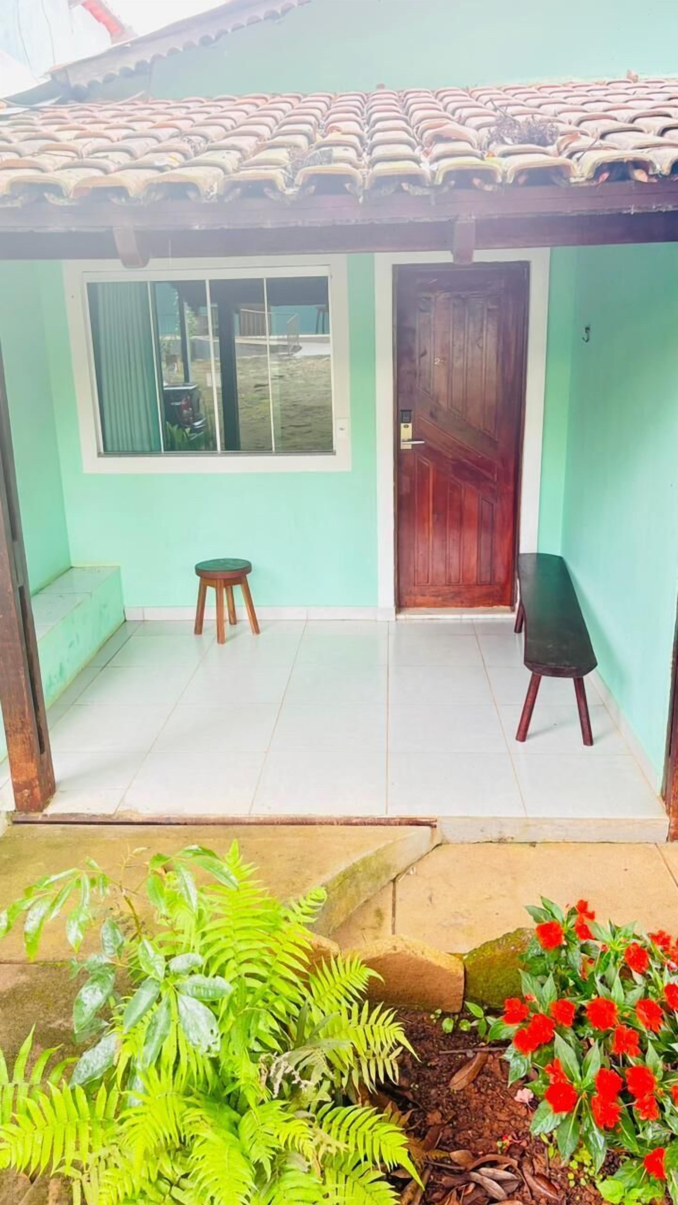 Una casita con techo de tejas, puerta de madera, ventana con cortinas, un taburete y una banca.
