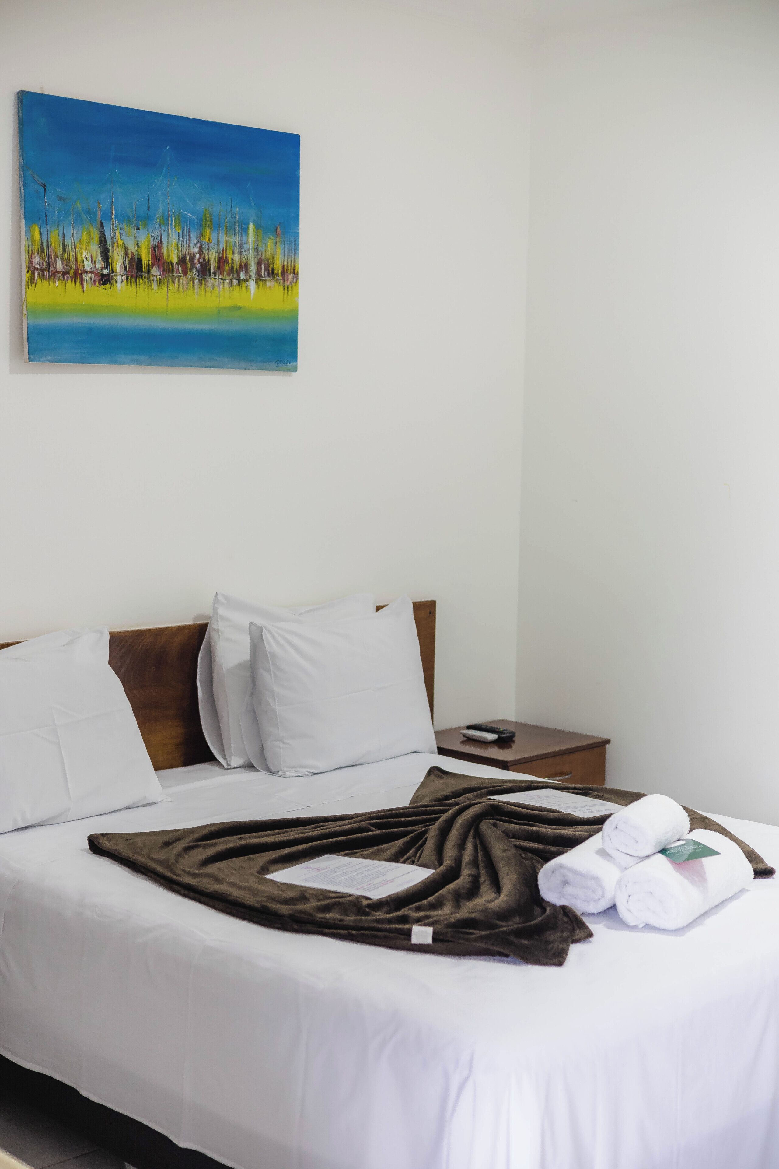 A hotel room with a bed, white linens, a brown throw blanket, a remote control, and a painting on the wall.