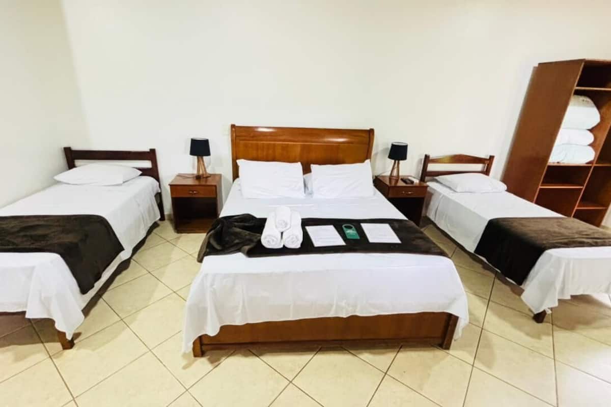 A hotel room with three single beds, a wooden wardrobe, and a small table with towels and a green card.