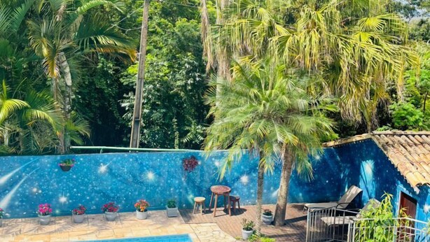 A swimming pool with a tiled deck, surrounded by lush greenery and a blue wall. - Chalés Casa Rosa (Alto Paraíso de Goiás)