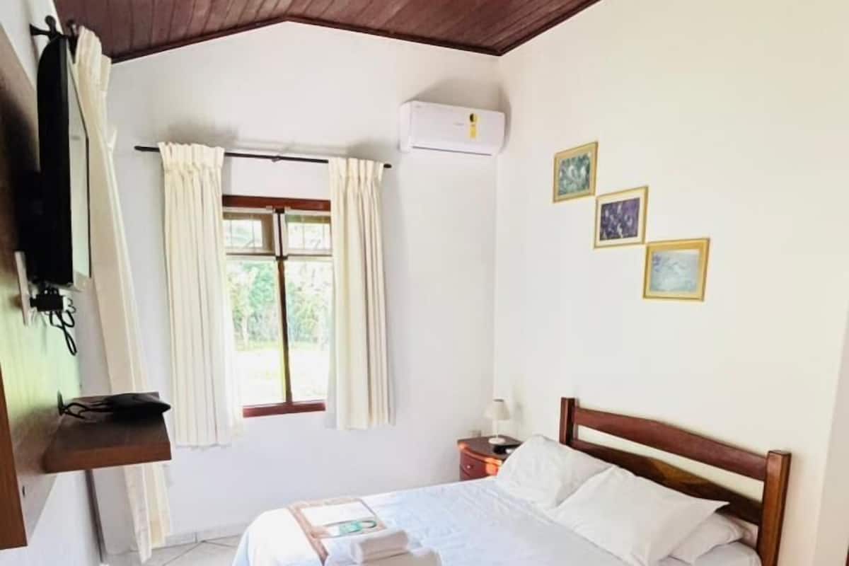 A hotel room with a wooden ceiling, a bed with white linens, a window with curtains, an air conditioning unit, and a small table with a paper.
