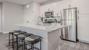 A modern kitchen with white cabinets, stainless steel appliances, and a central island with bar stools. - Landing | Beautiful 2BD, Pool (Tice)