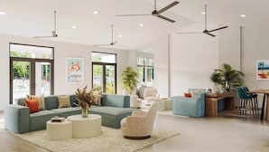 A spacious living room with a large sofa, a coffee table, and a dining area with a wooden table and chairs. There are ceiling fans, large windows, and a view of the outdoors. - Landing | Beautiful 2BD, Pool (Tice)