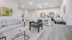Una sala de estar moderna con sofá, sillón y mesa de centro, comunicada con una cocina que tiene una isla central y un comedor con mesa y sillas.