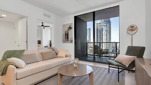 Una sala de estar moderna con un sofá, una mesa de centro y un sillón. Hay un ventanal con vistas a edificios altos.