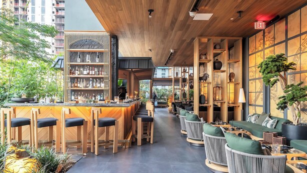 Zona de bar moderna con taburetes de madera, una barra con botellas y asientos con cojines verdes.