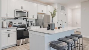 A modern kitchen with white cabinetry, stainless steel appliances, a central island with bar stools, and a vase with flowers. - Landing | Incredible 2BD, Pool (Tice)