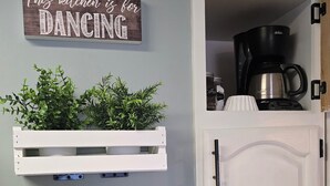 Uma cozinha com um letreiro que diz "Esta cozinha é para DANÇAR", prateleiras brancas suspensas com plantas e uma cafeteira.