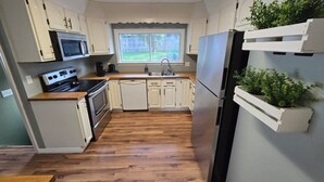 Cozinha compacta com armários brancos, forno embutido, micro-ondas, pia e geladeira. Há uma janela que oferece iluminação natural e um pequeno vaso de planta no balcão.