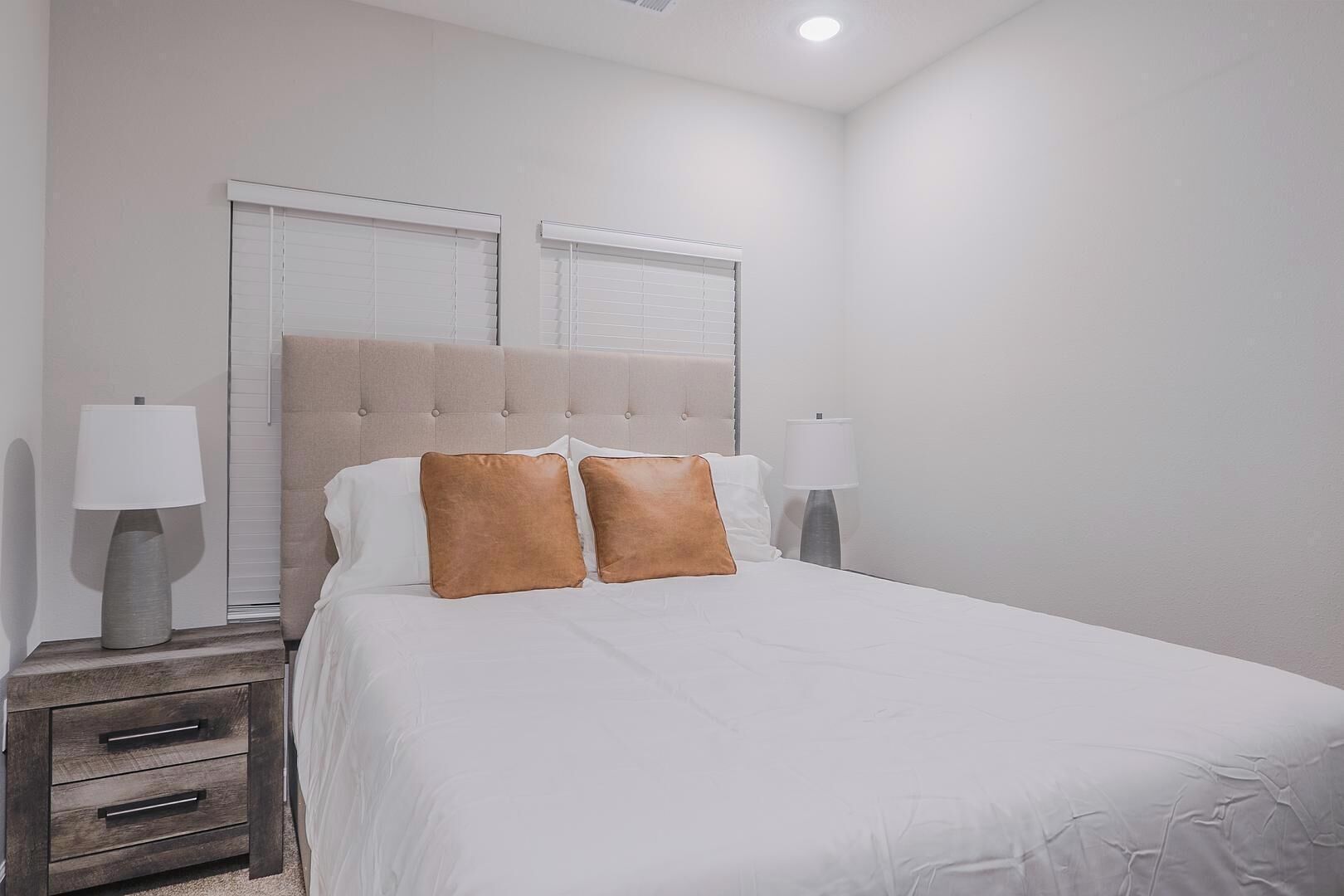 Un lit bien fait, avec du linge de lit blanc et deux oreillers marron, flanqué de deux tables de chevet avec des lampes, dans une pièce au design simple et épuré.