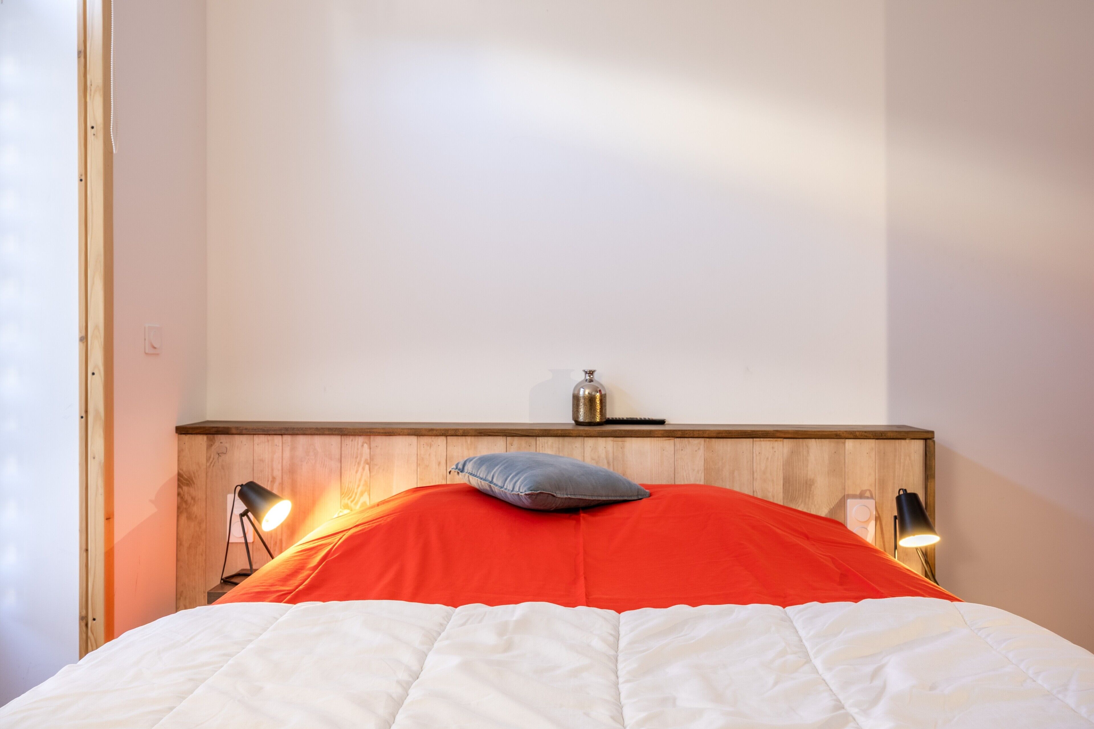 A bed with a red blanket, a white duvet, and a pillow. There are two bedside lamps and a small speaker on the wooden headboard.
