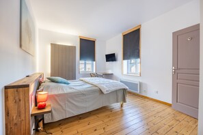 Une chambre à coucher comprenant un lit, une table de chevet avec une lampe, une armoire en bois, un téléviseur fixé au mur, des fenêtres et une porte.