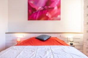 Ein ordentlich gemachtes Bett mit weißer und roter Decke, zwei Nachttischlampen und einem an der Wand hängenden Bild von einer Blume.