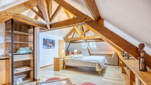 Quarto no estilo loft com vigas de madeira aparentes, cama, escrivaninha, cadeira e um banquinho vermelho.