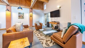 Uma sala de estar moderna com sofás de couro, uma mesa de centro de vidro, lareira, televisão e vigas de madeira no teto.