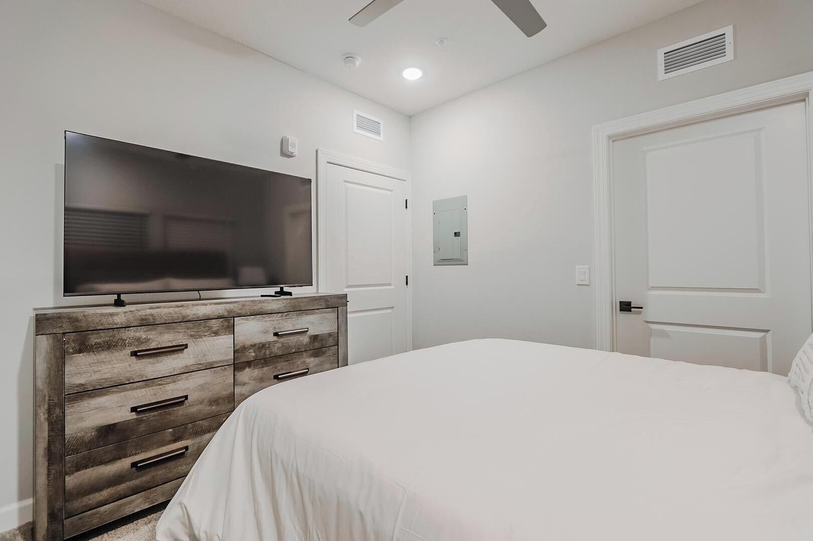 Un dormitorio con un televisor de pantalla plana grande sobre una cómoda de madera, una cama blanca y dos puertas blancas.