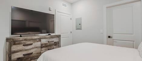 Un dormitorio con un televisor de pantalla plana grande sobre una cómoda de madera, una cama blanca y dos puertas blancas.