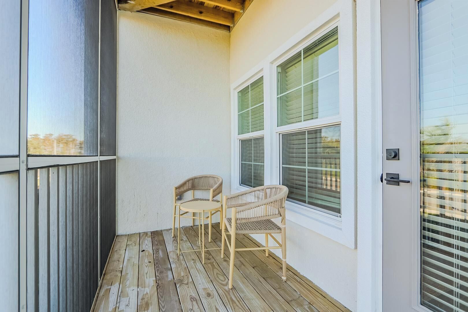 Un balcon doté d’un plancher en bois, de deux fauteuils en osier et d’une porte blanche avec une fenêtre.