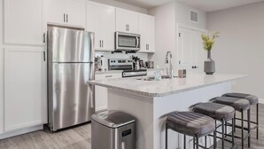 A modern kitchen with white cabinets, stainless steel appliances, a central island with bar stools, and a small trash bin. - Landing | Beautiful 2BD, Pool (Tice)