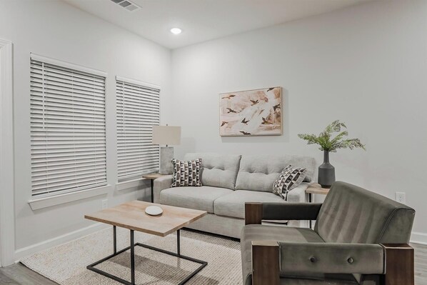 A living room with a grey sofa, a wooden coffee table, and a grey armchair. There is a painting on the wall and a vase with plants on a side table. - Landing | Amazing 2BD, Gym, Pool (Tice)